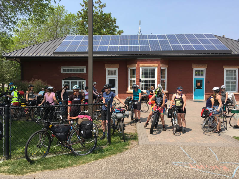A group a cyclists gathered around a red building, with solar panels on the roof