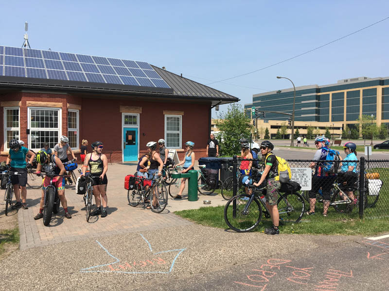 A group of cyclists in front of a red building with solar panels on the roof, with an office building background