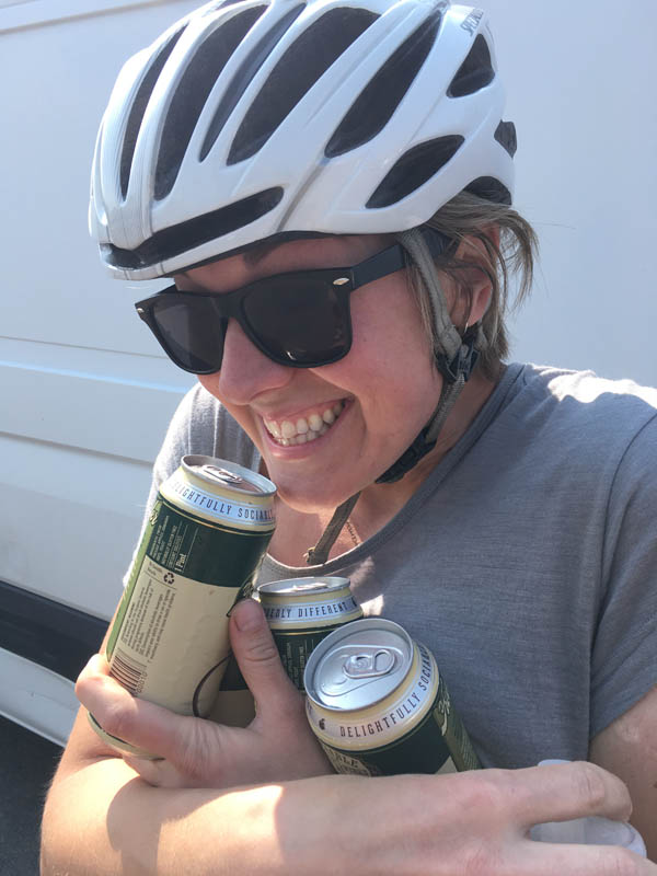 A smiling person wear a bike helmet and sunglasses, hugging 3 beer cans, with a white van in the background