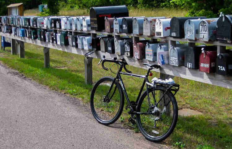 Left side view of a green Surly bike, parked along the side of a paved road, and leaning against a long row of mailboxes