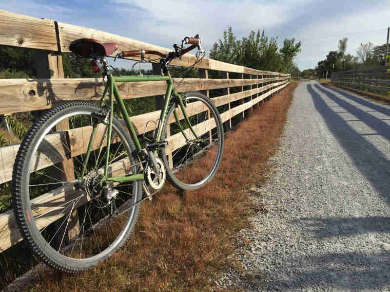 Rear view of a green bike, parked in the grass, leaning against a wood fence facing down a gravel road