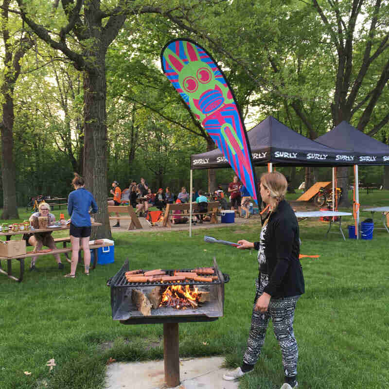 A person cooking at a park grill, with people, Surly canopies and trees in the background