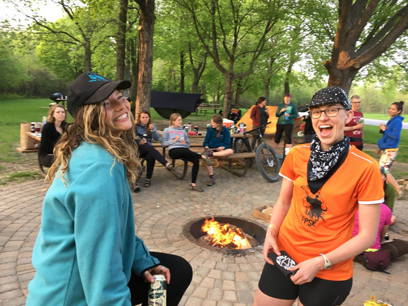 2 people posing for a picture in front of a patio fire pit, with people sitting around and trees in the background