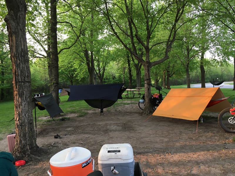 A dirt campsite inside a circle of trees with tents and hammocks attached
