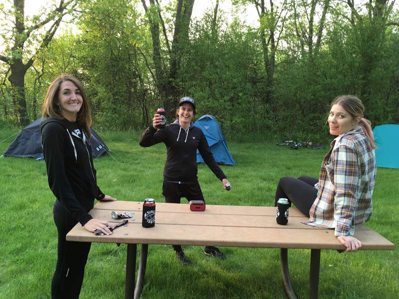 3 people gathered around a picnic table in a grass field with tents and trees in the background