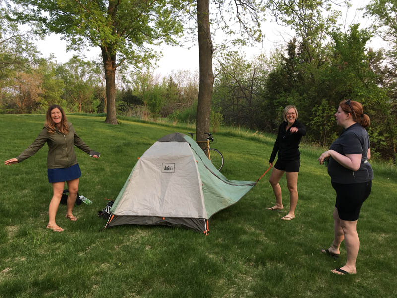 3 people putting up a tent in a grass field with trees in the background