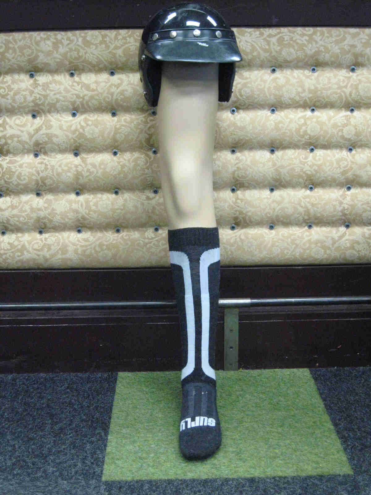Front, vertical view of a mannequin leg, wearing a Surly Tall Sock and a helmet on top, leaning against a padded wall