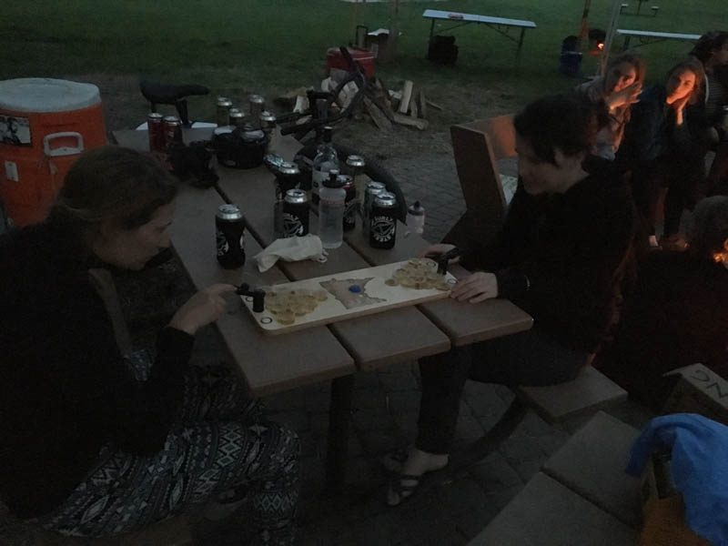 2 people sitting on a picnic table, playing a board game, with people around a campfire in the background, at night