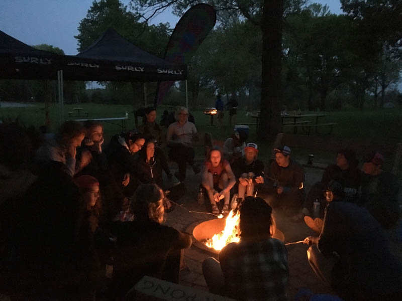 Group of people gathered around a campfire at night, with Surly canopies in the background