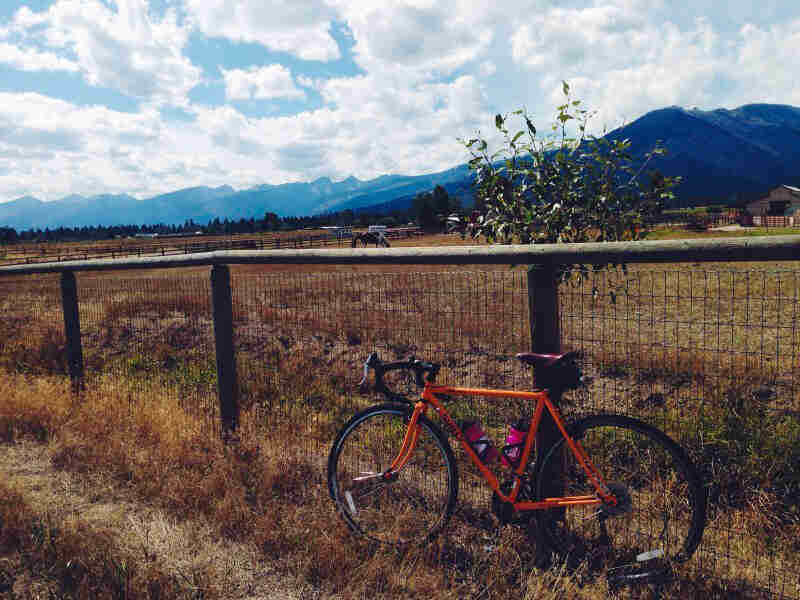 Left side view of an orange Surly bike, leaning on a fence with a horse in a pasture behind, and mountains in background
