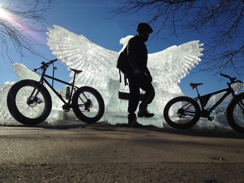 Shadowy view of a cyclist, standing between two Surly fat bikes facing opposite directions, in front of an ice sculpture
