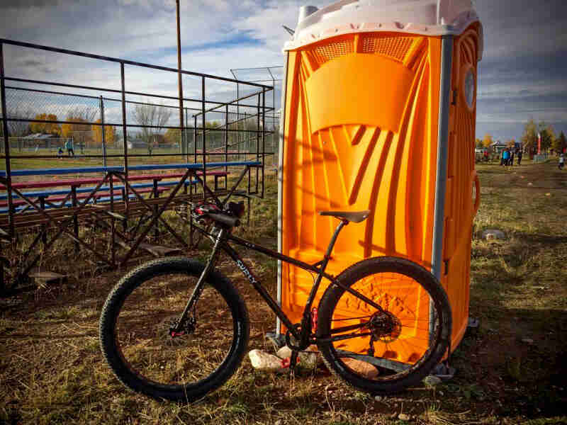 Left side view of a Surly bike, parked in front of an orange porta-potty, with a baseball field in the background