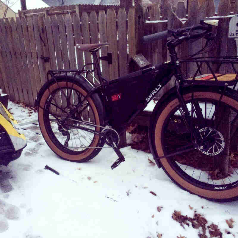 Right side view of a black Surly fat bike with racks and a frame bag, parked on snow next to a wood fence