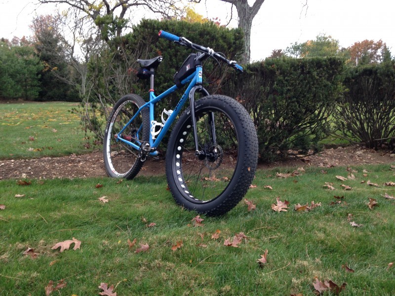 Front, right side view of a blue Surly fat bike, parked on a grass field, next to a hedge bush and tree