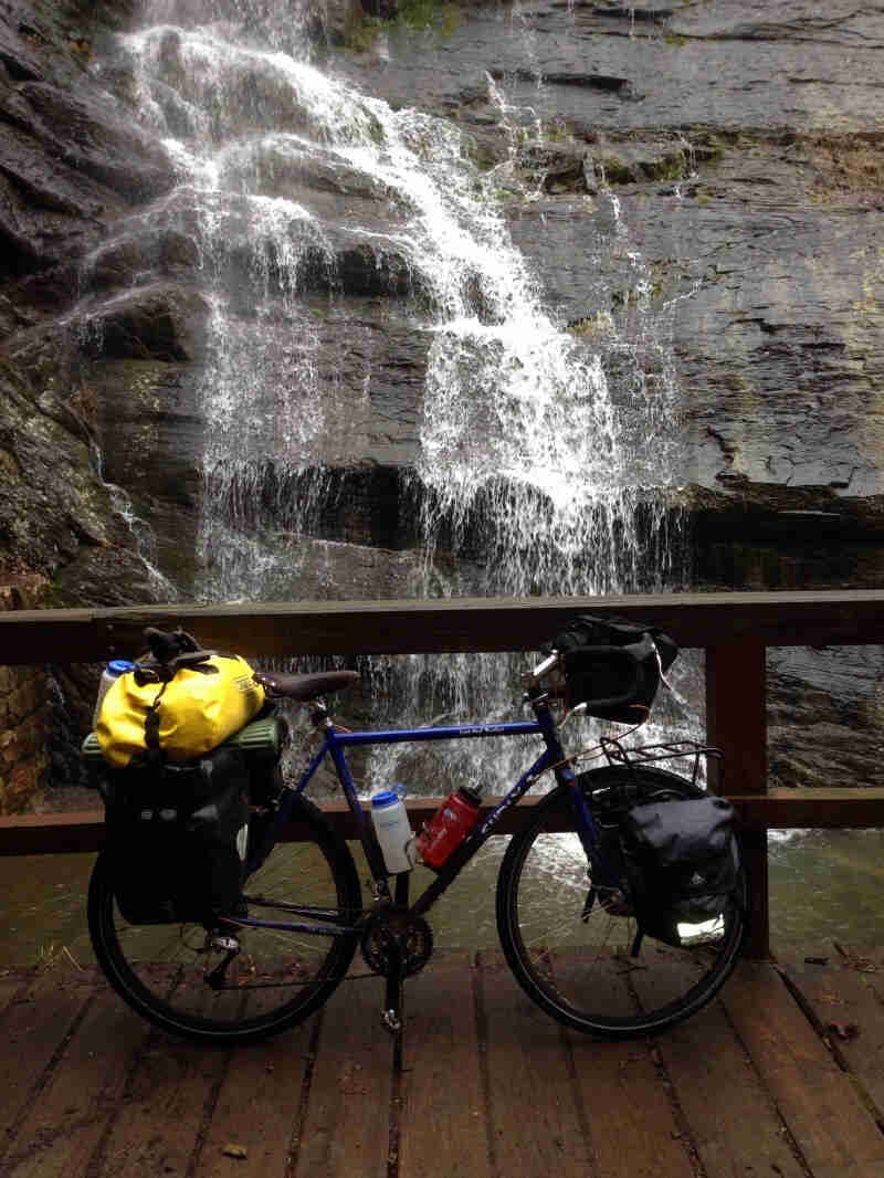 Right side view of a blue Surly Long Haul Trucker bike with gear, leaning on a rail of a bridge with a waterfall behind
