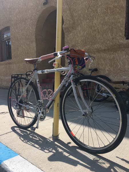 Right side view of a Surly Cross-Check bike, gray, chained to a yellow post, on a sidewalk, in front of a building