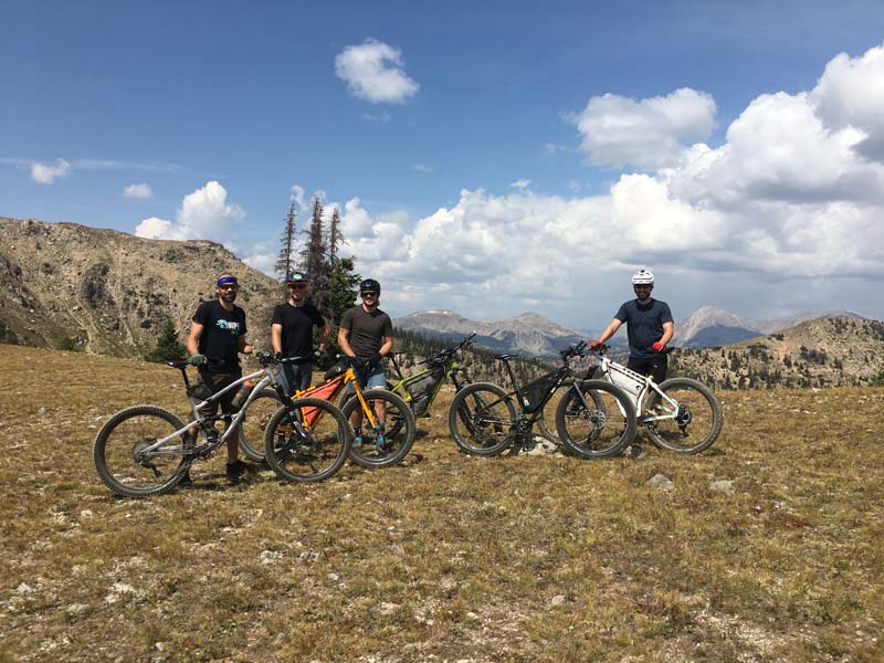 Group of cyclists standing with their bikes, on a grassy field, with mountains in the background
