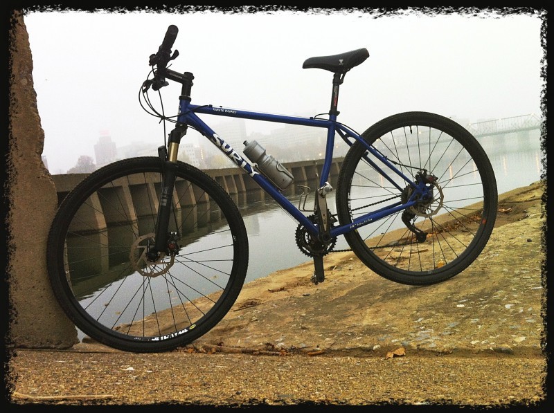 Left side view of a blue Surly Karate Monkey bike, parked on a concrete slab, above water with a cement pier behind it