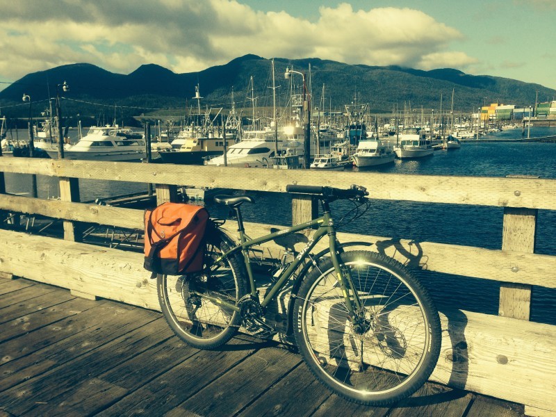 Right side view of a green Surly bike with a rear bag, leaning on a rail of a wood dock, with boat in a marina behind it