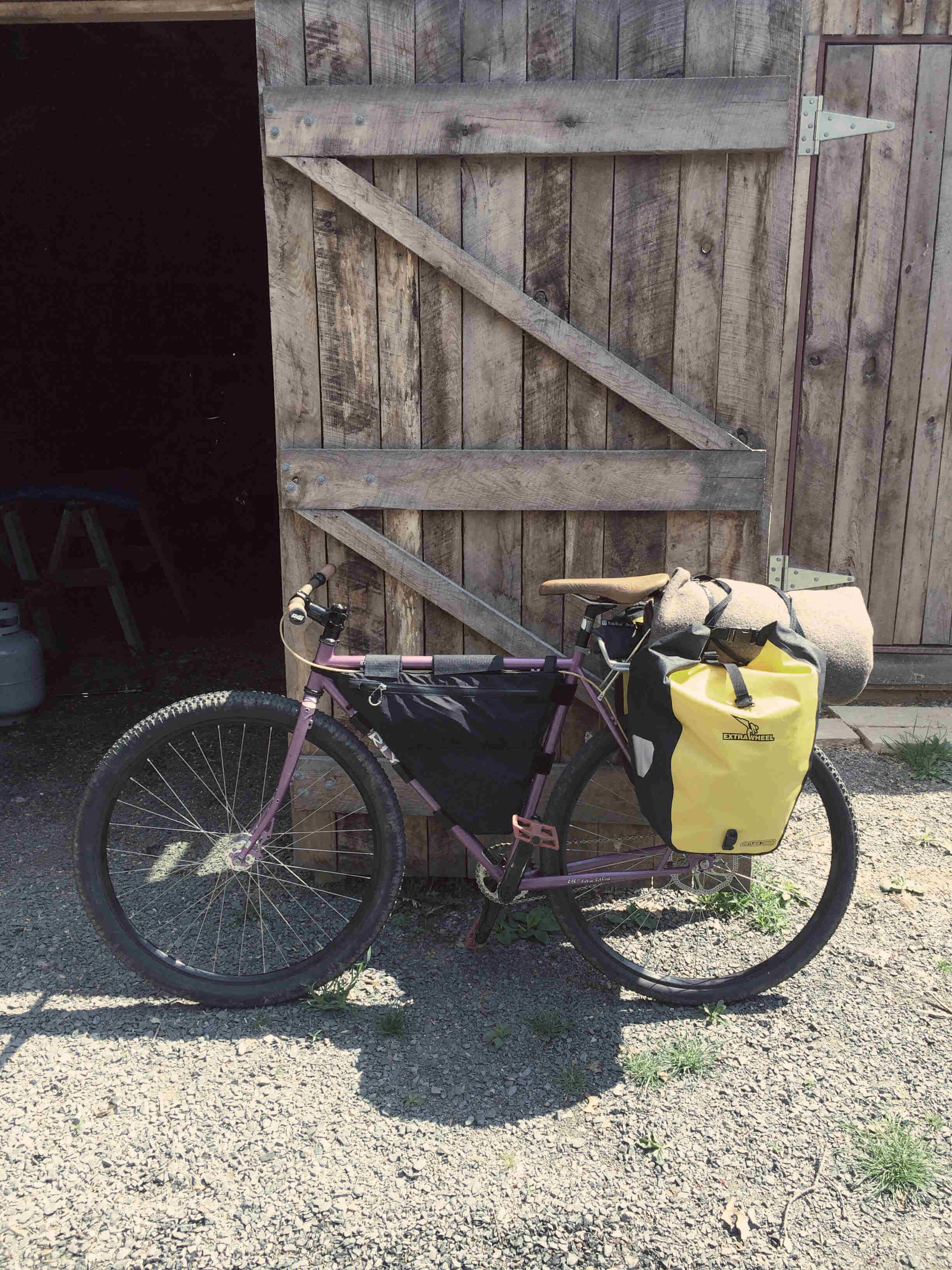 Right side view of a lavender color Surly bike with gear packs, parked against the door of a barn on gravel