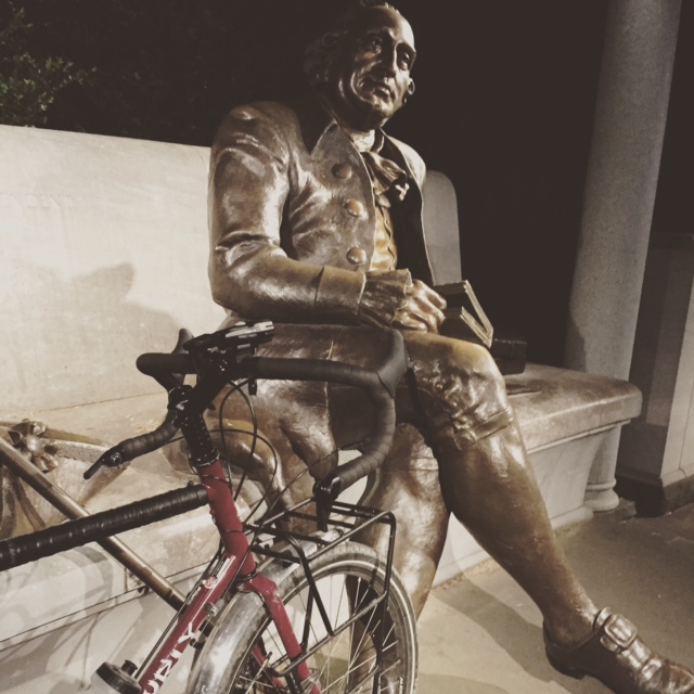 Cropped, right side view of the frontend of a red Surly bike, in front a bronze statue of a person sitting on a bench