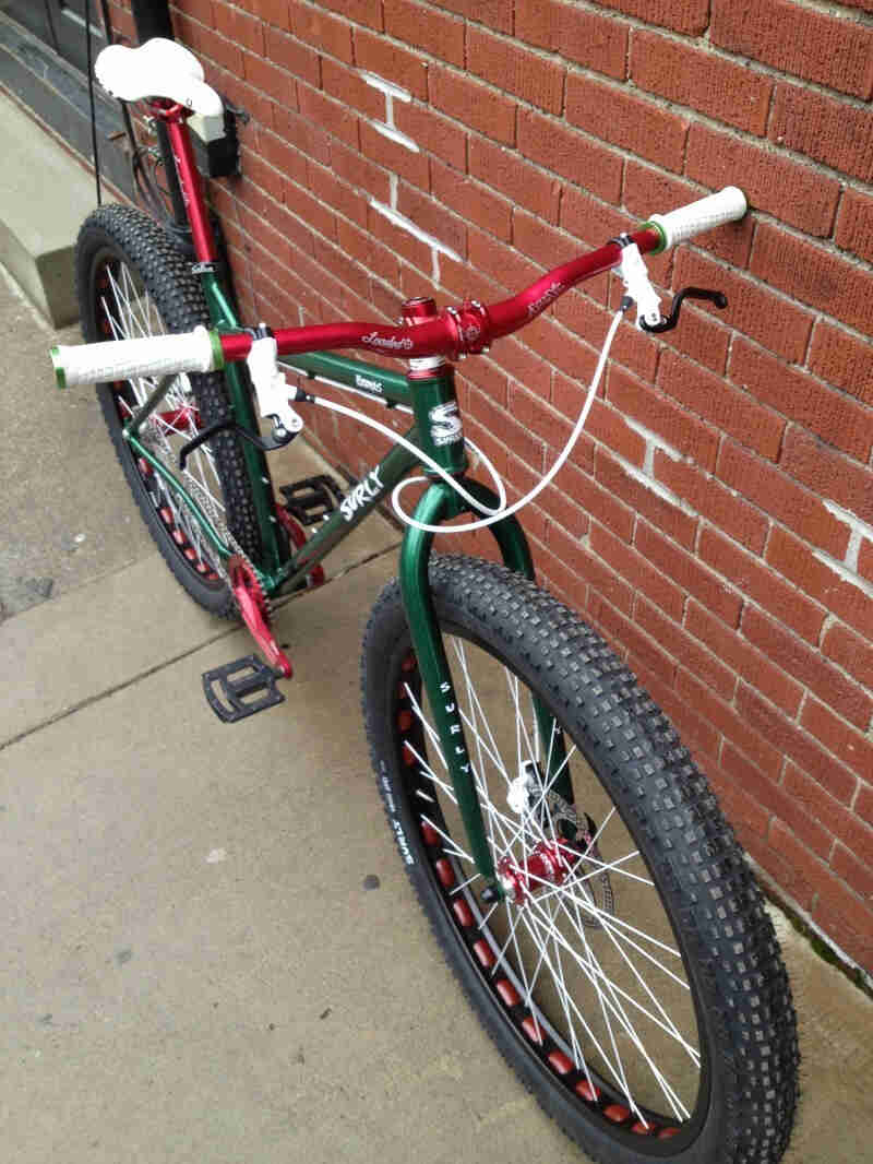 Downward, front, right side view of a green Surly Krampus bike, leaning against a brick wall, on a sidewalk