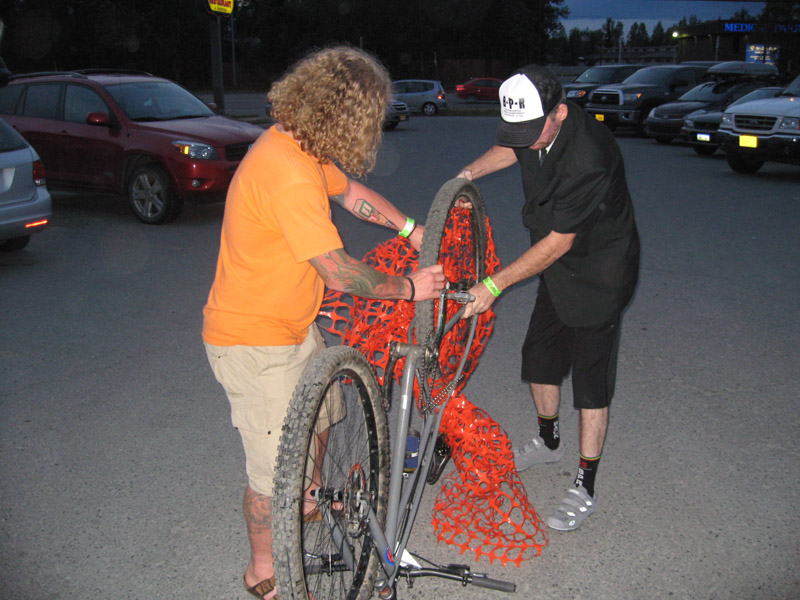 Side view of two people, untangling orange snow fence from the rear wheel of a bike, at night on a parking lot