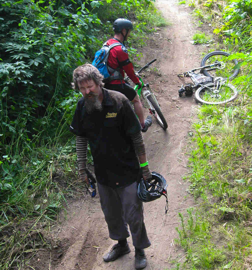 Front view of a cyclist standing on a dirt trail in the woods, with their bike laying behind, next to another cyclist