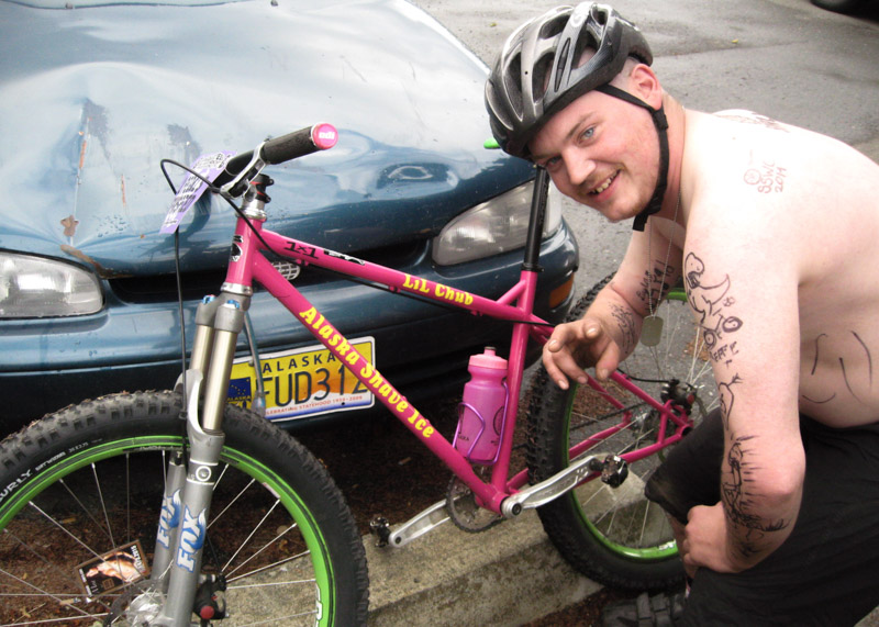 Left side view of a pink Surly 1x1 bike leaning on the front of a car, with a cyclist kneeling down by the rear wheel