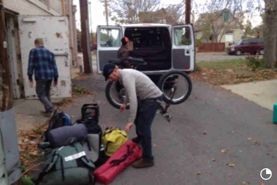 A person standing over a pile of biking packs, with a bike and a van in the background