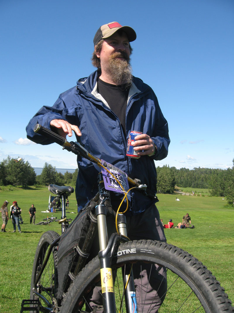 Front view of a cyclist, standing with a Surly bike, with a can in hand, and a grass field with people in the background