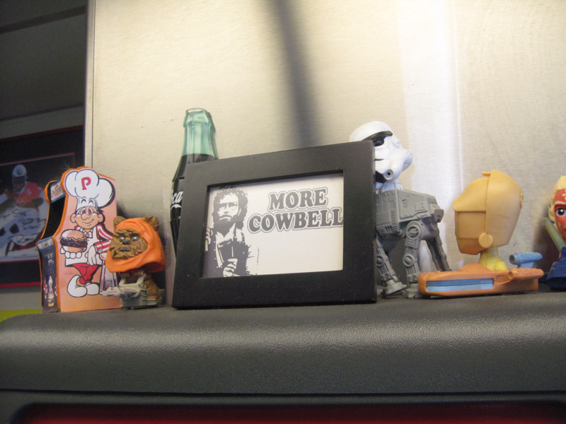 A framed illustration of a Saturday Night Live tv character, with the words More Cowbell, on a shelf with toy figures
