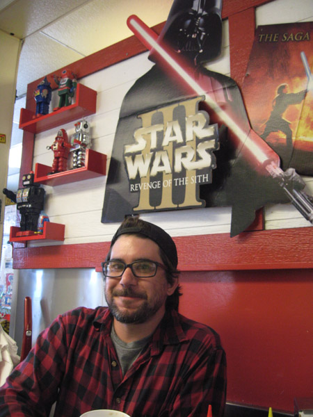 Front view of a person wearing glasses, sitting at a table with a Star Wars movie poster on the wall behind them