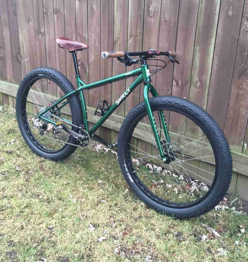 Right side view of a green Surly Krampus bike, parked on grass, against a wood fence wall
