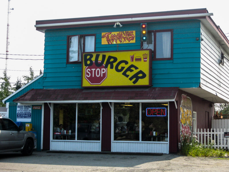 Front of a teal and brown restaurant building with an neon OPEN sign in the window, and a Burger Stop sign