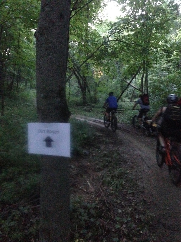 Rear view of cyclists riding their bikes on a muddy trail in the woods, with a Dirt Burger flyer on a tree to the left