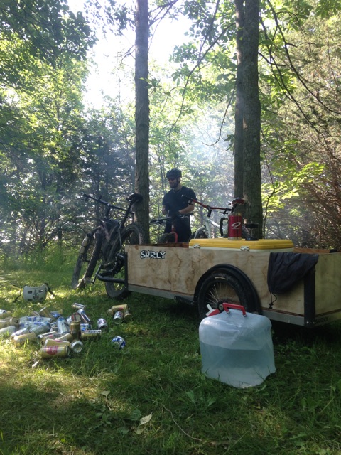 Beer cans and a water jug, laying in front of a bike with a trailer, and a person standing in the background