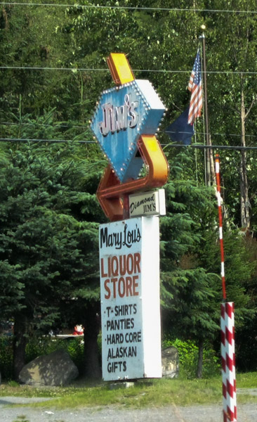 A liquor store sign, on a roadside, with pine trees behind it