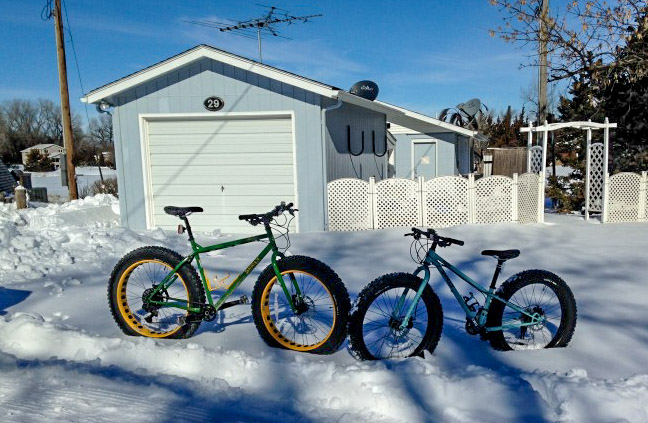 Two Surly fat bikes facing each other, in deep snow, with a yard and garage in the background