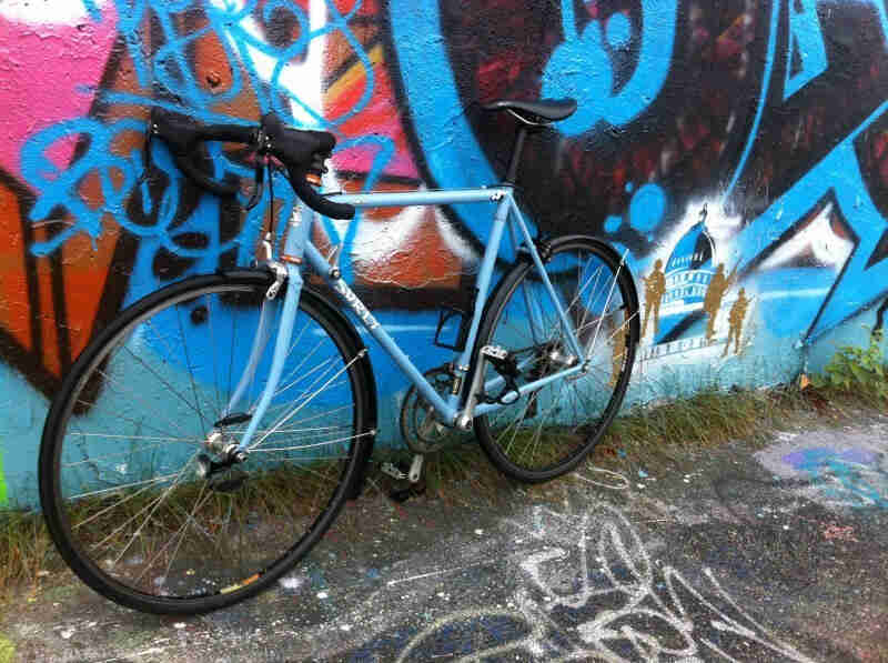 Left side view of a light blue Surly bike, leaning against a cement wall that's painted with graffiti