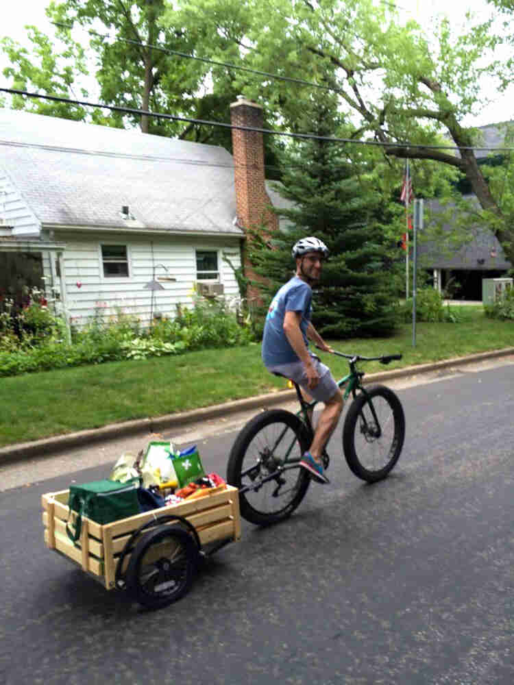 Rear, right side view of a cyclist on a green bike with trailer, riding down a street, with a house in the background