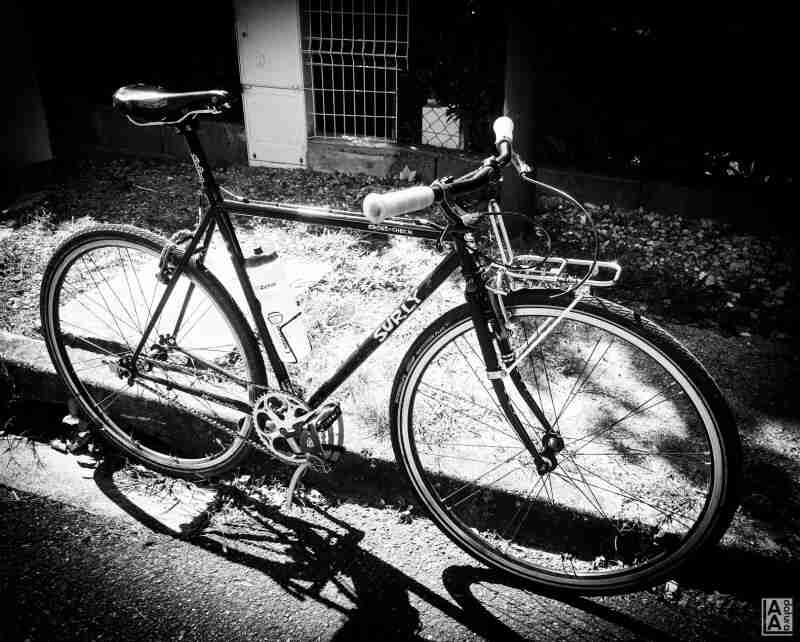 Black & white, right side view of a Surly Cross Check bike, parked against a curb with a wire fence in the background