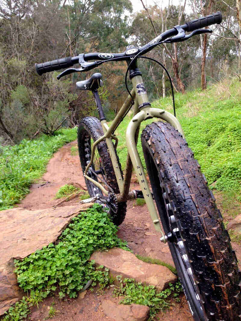 Front, close up view of an olive green Surly fat bike, facing up a muddy trail, on a hill in the woods