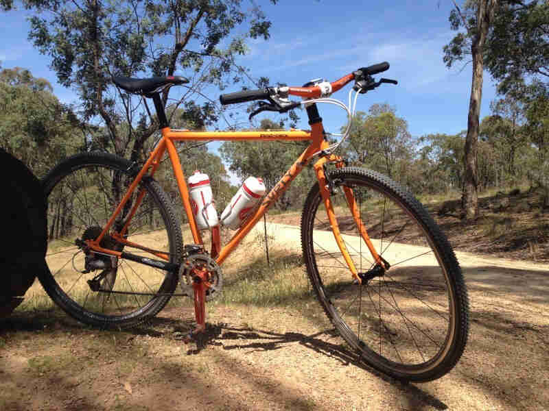 Right side view of an orange Surly Cross Check bike, parked in grass, on the side of a gravel road