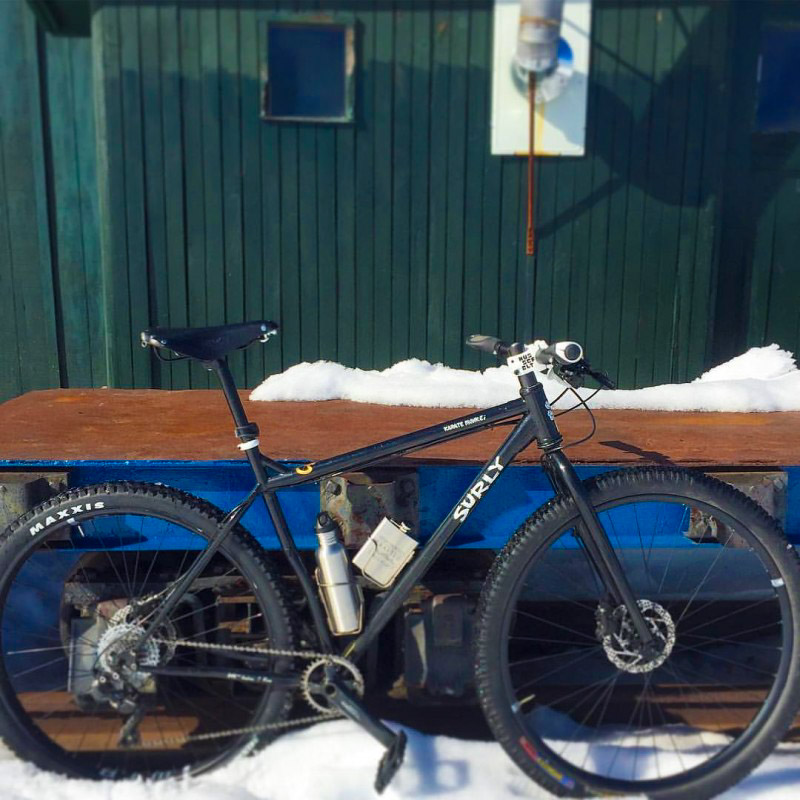 Right side of a black Surly Karate Monkey bike, parked on snow and leaning on a table, with a green building behind