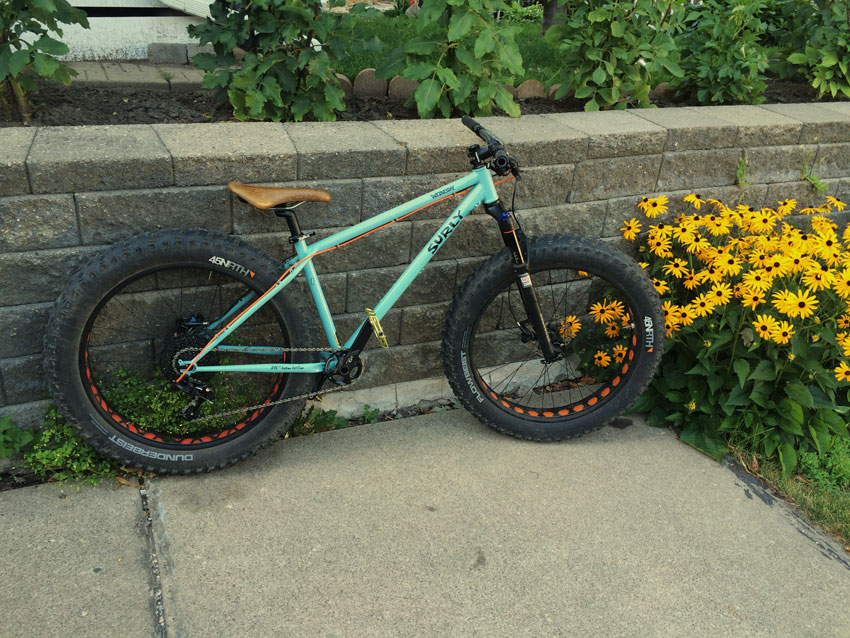 Right side view of a mint Surly Wednesday fat bike, leaning against a block wall on a patio, next to yellow flowers