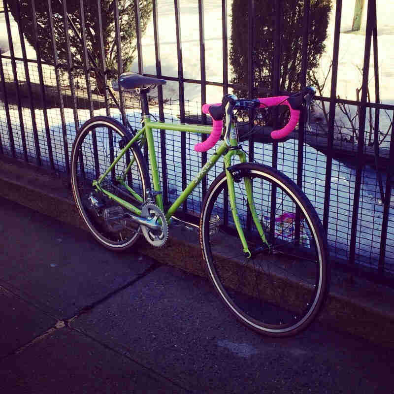Right side view of a lime green Surly bike with pink handlebar, parked on a sidewalk, against a steel rod fence