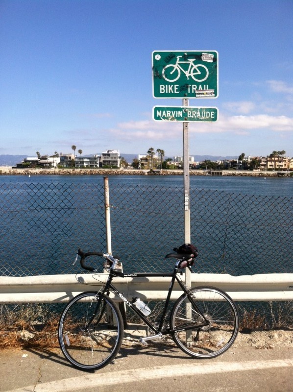 Left side view of a black Surly Long Haul Trucker bike, leaning on a road guardrail, with a bay and buildings behind it