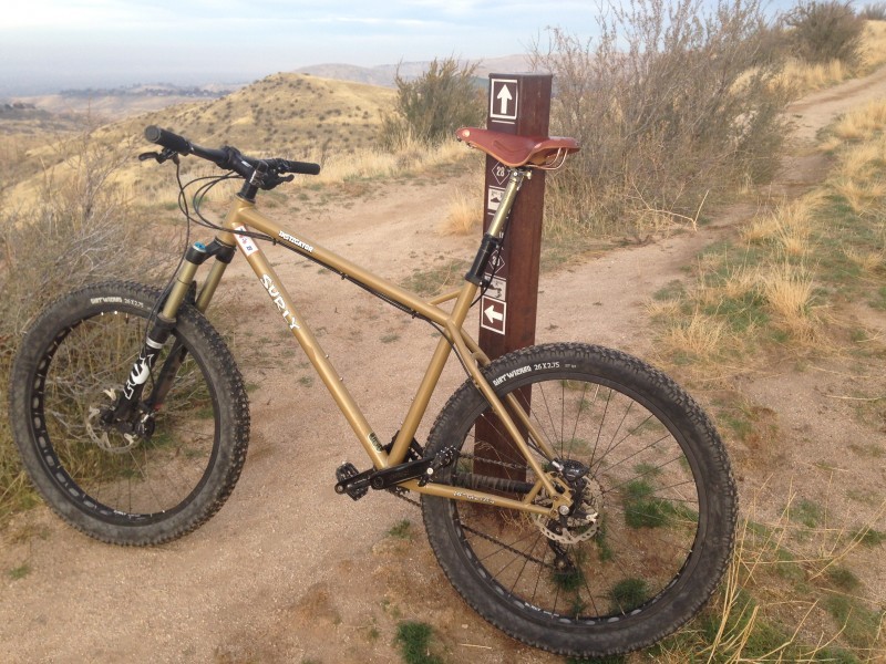 Left side view of a mustard yellow Surly Instigator bike, leaning on a post, at a dirt trail in the grassy desert hills