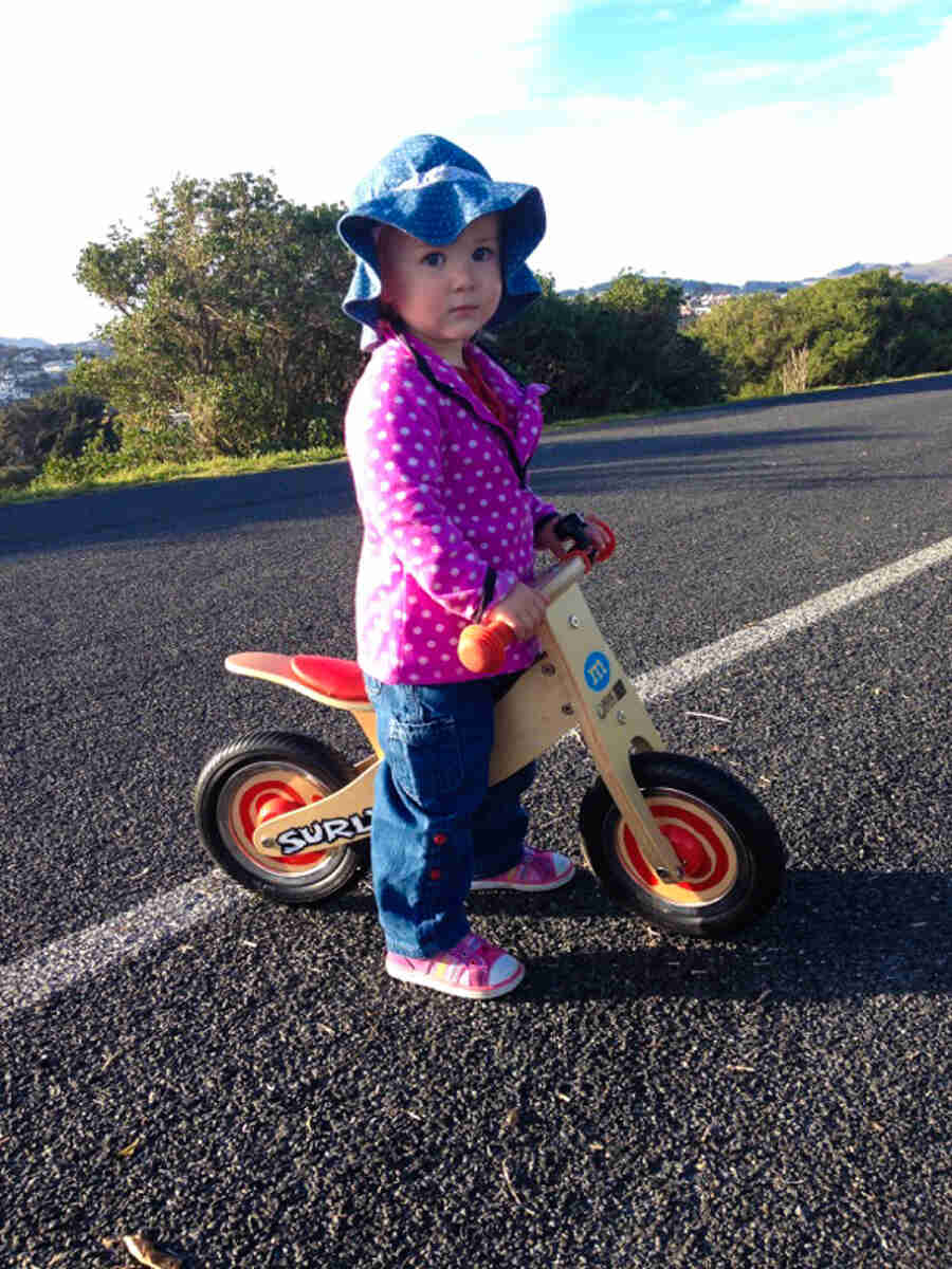 A small child standing over a plastic kids bike with a Surly sticker on the back, parked on a paved trail 
